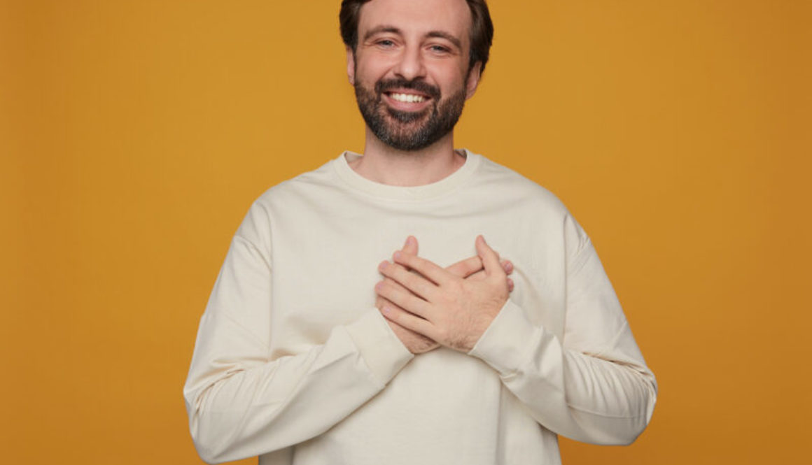 indoor portrait of handsome bearded male posing over orange background keeps his palm on heart, eye closed and smiles broadly